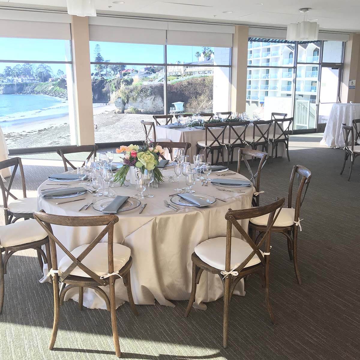 A dining area with a round table set for a meal, featuring a white tablecloth, glassware, silverware, and a flower arrangement, with a scenic view of the ocean outside.